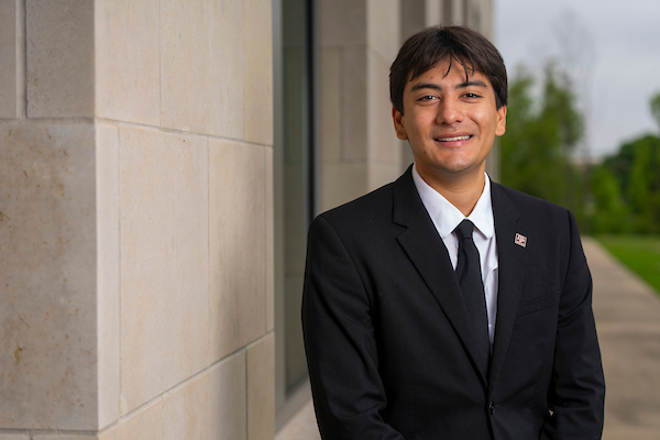 A male Hispanic student in a suit posing by a building