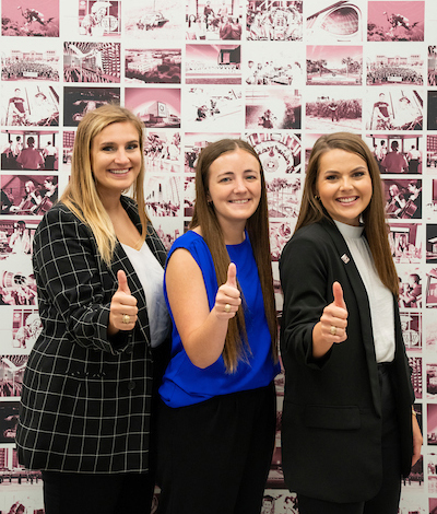 three students giving a thumbs-up in front of a wall of art
