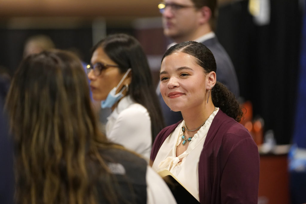 Close-up of woman dressed professionally and smiling at another woman in a crowd.