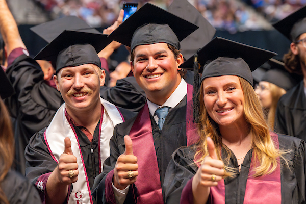 Three students in graduation robes giving a thumbs up